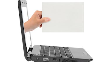 laptop stojący bokiem, z którego wyłania się dłoń trzymająca białą kartkę